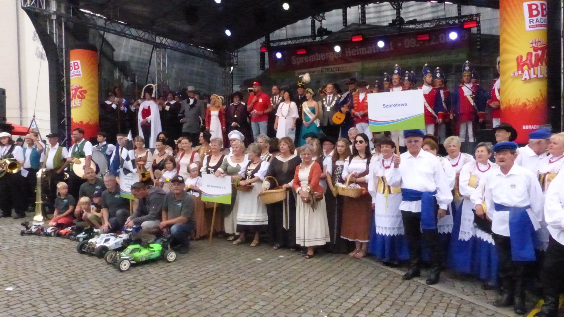 Gruppenfoto auf der Bühne des Spremberger Heimatfests mit Teilnehmern in traditionellen Trachten, Musikern, historischen Uniformen und Vereinsmitgliedern vor dem BB Radio Bühnenbanner.